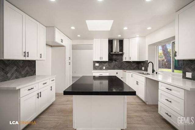 kitchen featuring a skylight, white cabinetry, sink, wall chimney range hood, and a kitchen island