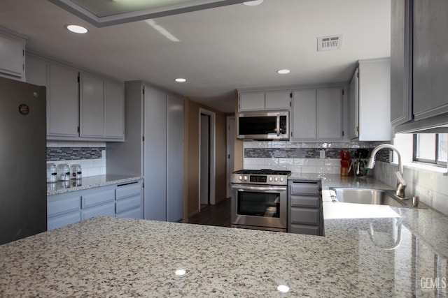 kitchen with sink, light stone counters, backsplash, gray cabinets, and appliances with stainless steel finishes