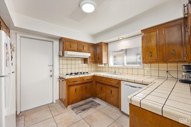 kitchen featuring tile countertops, under cabinet range hood, white appliances, backsplash, and brown cabinetry