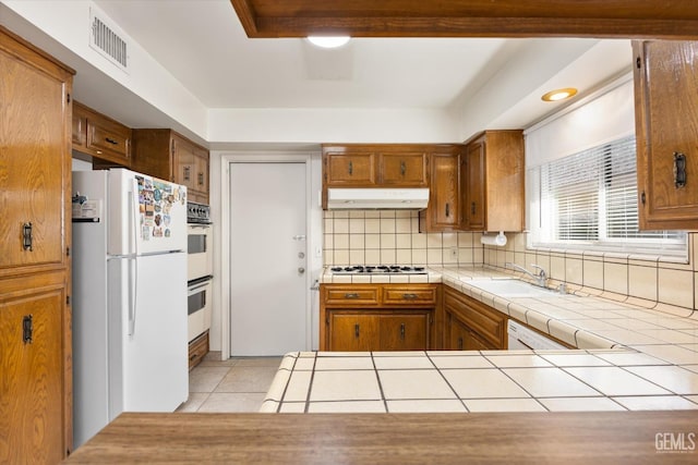 kitchen featuring white appliances, tile countertops, brown cabinets, under cabinet range hood, and a sink