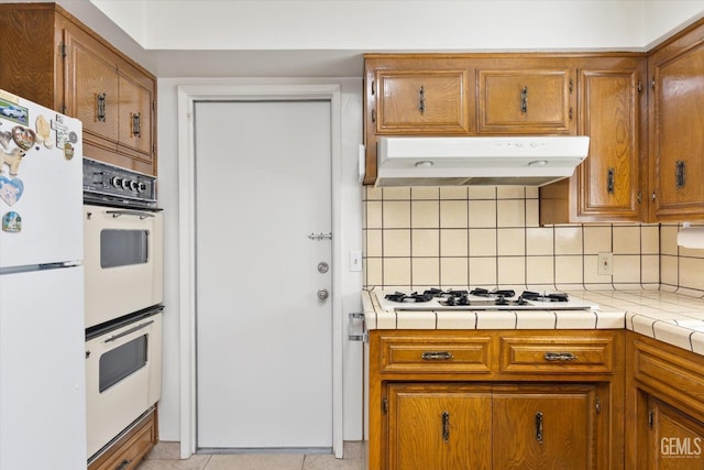 kitchen with white appliances, under cabinet range hood, brown cabinets, and backsplash