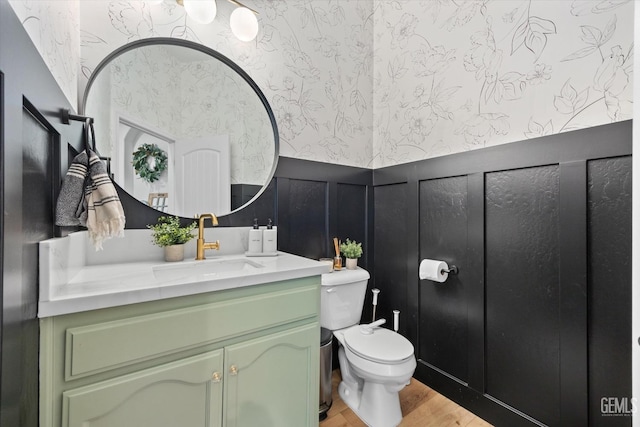 bathroom with wallpapered walls, wainscoting, toilet, wood finished floors, and vanity