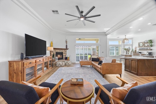 living room with light wood finished floors, visible vents, ornamental molding, ceiling fan, and a stone fireplace