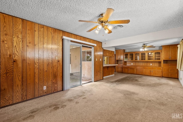 unfurnished living room with visible vents, wood walls, light colored carpet, a textured ceiling, and a ceiling fan