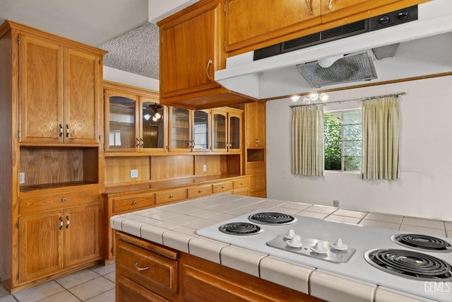 kitchen with white electric stovetop, tile countertops, light tile patterned floors, under cabinet range hood, and glass insert cabinets