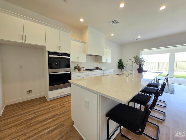 kitchen with a kitchen island with sink, a sink, white cabinetry, stainless steel double oven, and gas stovetop