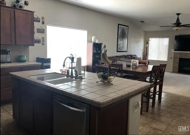 kitchen with a tile fireplace, a center island with sink, sink, ceiling fan, and tile counters