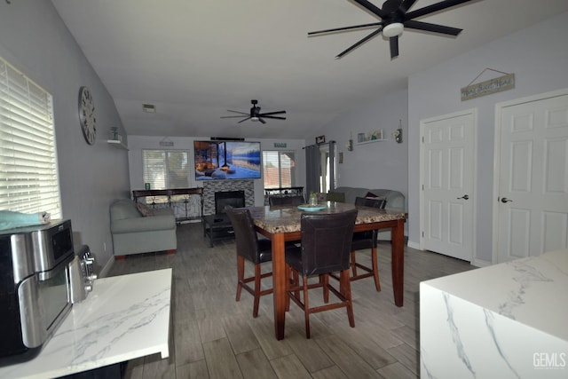 dining area with ceiling fan, a stone fireplace, and dark wood-type flooring