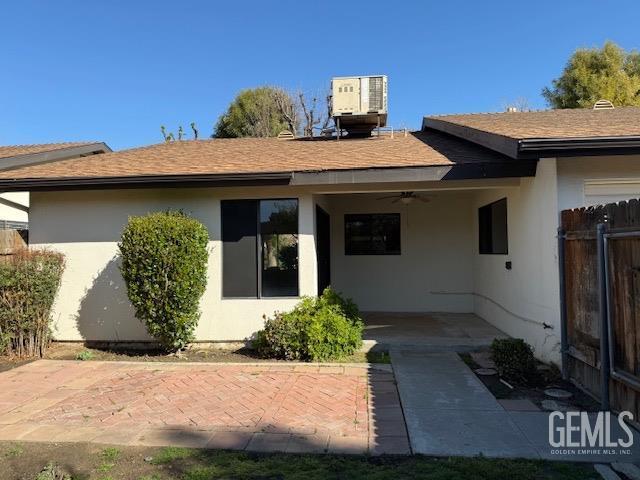 back of house with cooling unit, a ceiling fan, fence, roof with shingles, and stucco siding