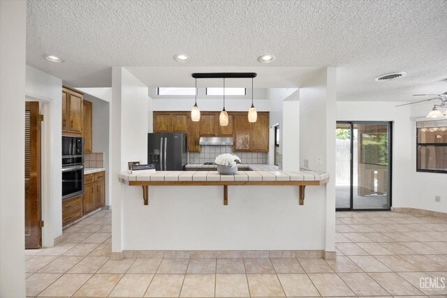 9408 Wandering Oak Dr, Bakersfield, CA 93311 - kitchen featuring stainless steel appliances, tile counters, brown cabinets, under cabinet range hood, and a textured ceiling