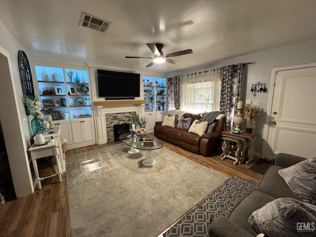 living room with visible vents, ceiling fan, wood finished floors, a stone fireplace, and built in shelves