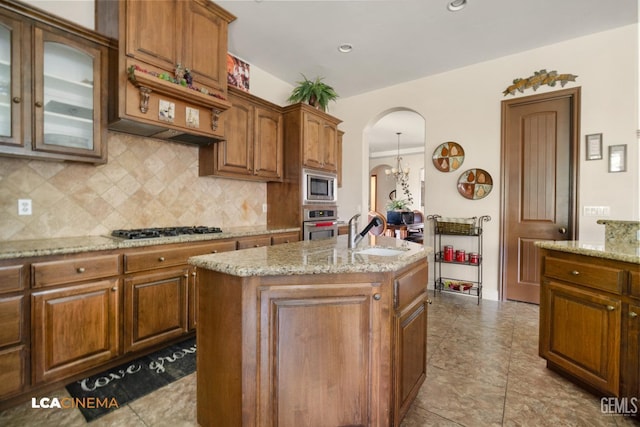 kitchen featuring a center island with sink, sink, decorative backsplash, light stone countertops, and appliances with stainless steel finishes