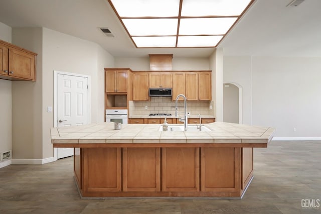 kitchen featuring decorative backsplash, tile countertops, oven, and a center island with sink