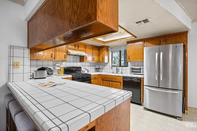 kitchen with tile counters, visible vents, a peninsula, under cabinet range hood, and black appliances