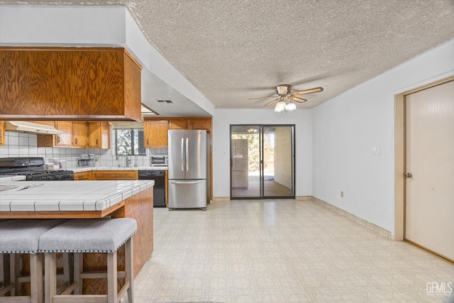 kitchen featuring tile countertops, light floors, decorative backsplash, black appliances, and brown cabinetry