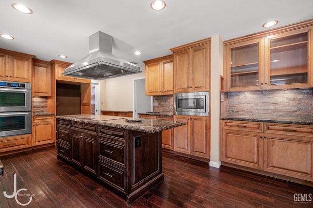 kitchen featuring stainless steel appliances, a center island, dark stone counters, island exhaust hood, and glass insert cabinets