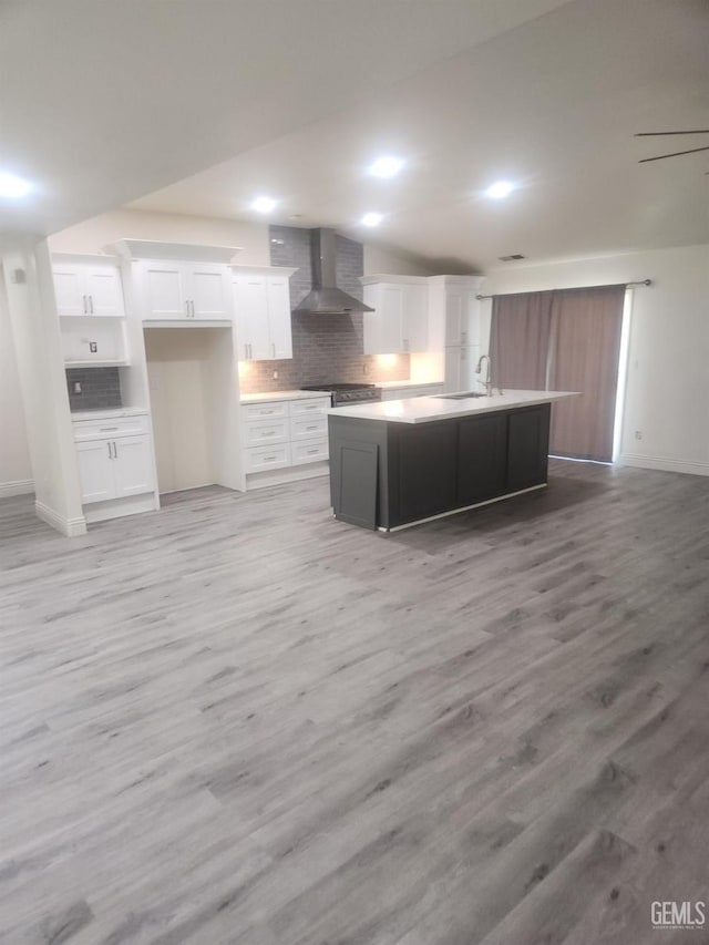 kitchen with wall chimney range hood, sink, white cabinetry, tasteful backsplash, and a center island with sink