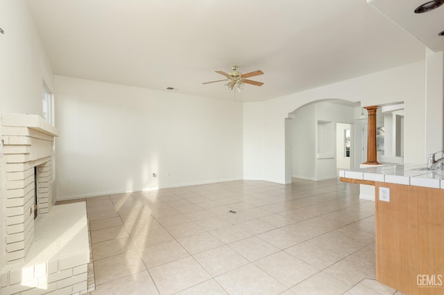 unfurnished living room featuring light tile patterned floors, arched walkways, a brick fireplace, and a ceiling fan