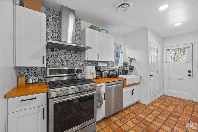 kitchen with stainless steel appliances, butcher block countertops, white cabinetry, and wall chimney exhaust hood