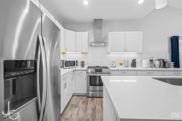 kitchen with wall chimney range hood, white cabinetry, light wood-style flooring, stainless steel appliances, and light countertops