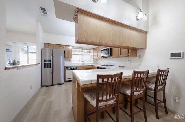 kitchen with tile counters, stainless steel appliances, light hardwood / wood-style flooring, kitchen peninsula, and a kitchen bar