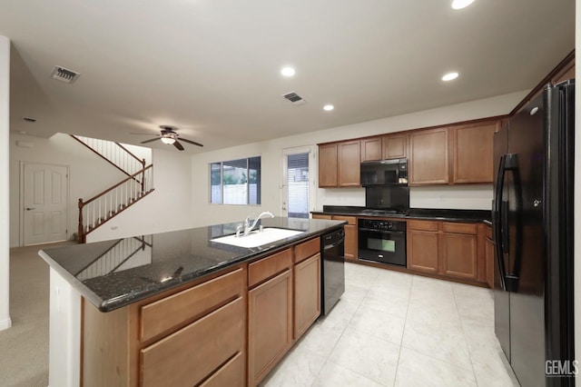 kitchen with a kitchen island with sink, black appliances, sink, ceiling fan, and dark stone countertops