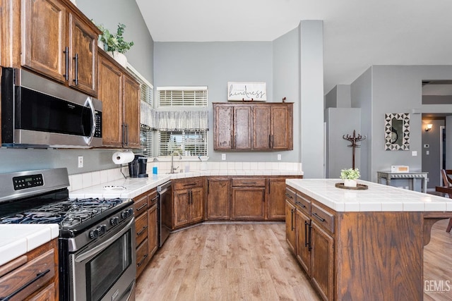 kitchen featuring tile countertops, stainless steel appliances, light wood-style flooring, a sink, and a kitchen island