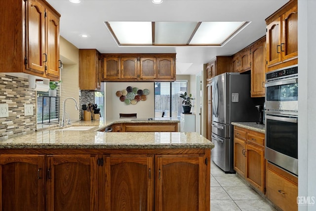 kitchen with appliances with stainless steel finishes, a peninsula, light stone countertops, a sink, and light tile patterned flooring