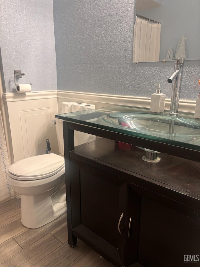 half bath with a textured wall, toilet, wainscoting, vanity, and wood finished floors