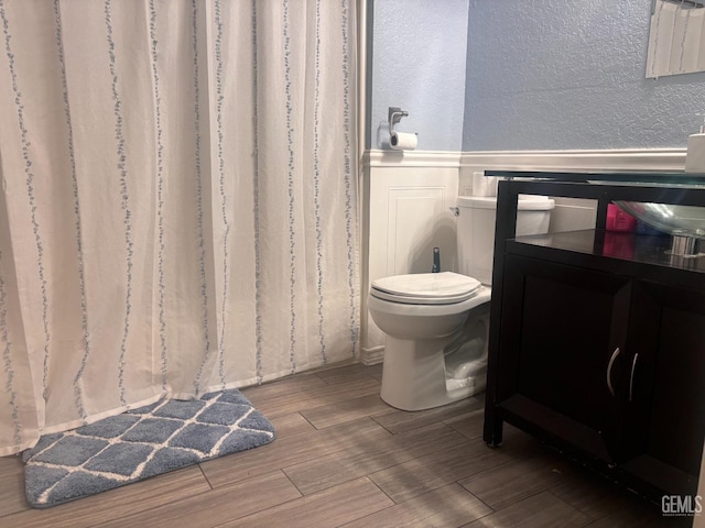 bathroom with wood finish floors, a wainscoted wall, a textured wall, a decorative wall, and toilet