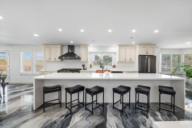 kitchen with recessed lighting, freestanding refrigerator, light countertops, cream cabinets, and wall chimney exhaust hood