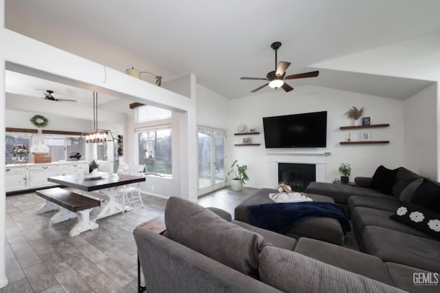 living area featuring a glass covered fireplace, wood finished floors, ceiling fan with notable chandelier, and vaulted ceiling