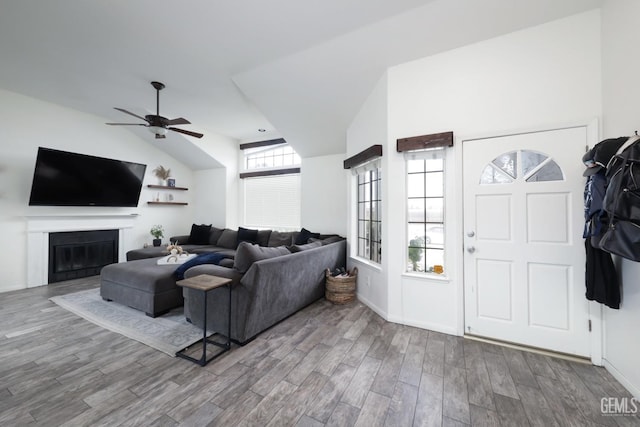 living area with baseboards, a glass covered fireplace, vaulted ceiling, wood finished floors, and a ceiling fan