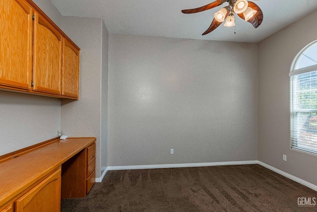 unfurnished office with a ceiling fan, dark colored carpet, built in desk, and baseboards