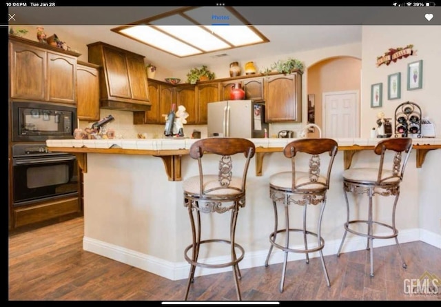 kitchen with dark wood-type flooring, a breakfast bar, tasteful backsplash, tile counters, and black appliances