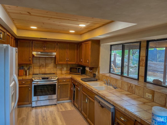 kitchen featuring appliances with stainless steel finishes, tile counters, under cabinet range hood, a raised ceiling, and a sink