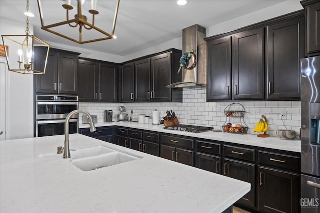 kitchen featuring wall chimney range hood, a sink, decorative backsplash, a chandelier, and stainless steel appliances