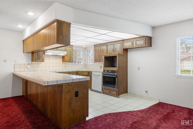 kitchen featuring a peninsula, white dishwasher, oven, decorative backsplash, and light carpet