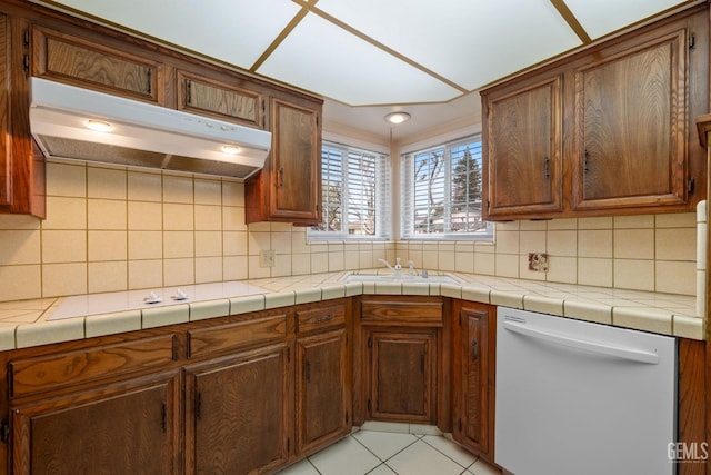 kitchen featuring light tile patterned flooring, a sink, under cabinet range hood, dishwasher, and tasteful backsplash