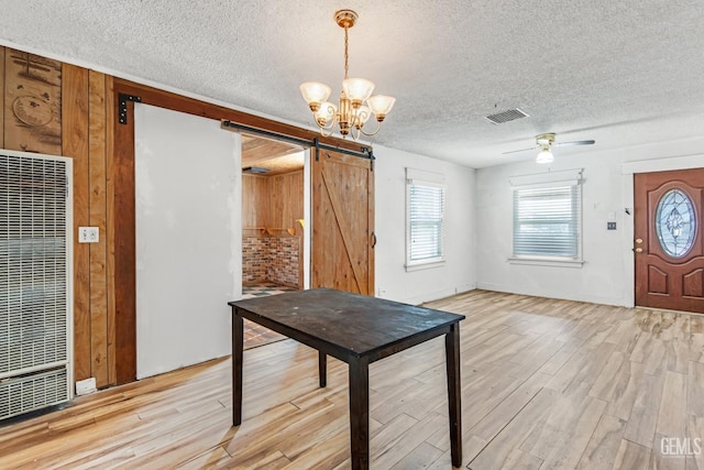 unfurnished dining area with ceiling fan with notable chandelier, a barn door, a textured ceiling, and light wood-type flooring