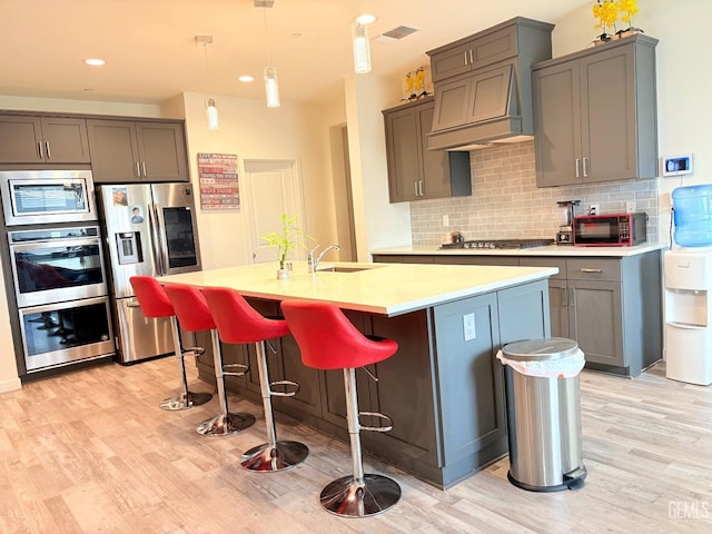 kitchen with appliances with stainless steel finishes, decorative backsplash, light hardwood / wood-style floors, hanging light fixtures, and a center island with sink