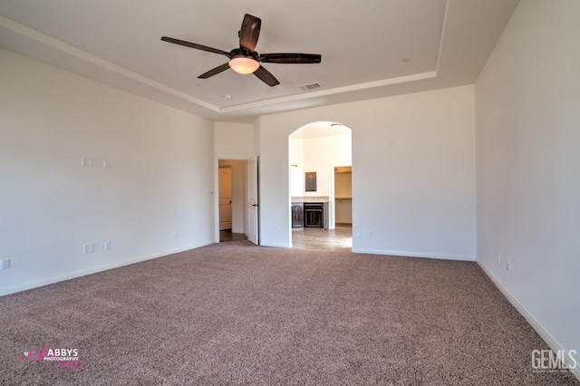 empty room with arched walkways, carpet floors, ceiling fan, and a raised ceiling