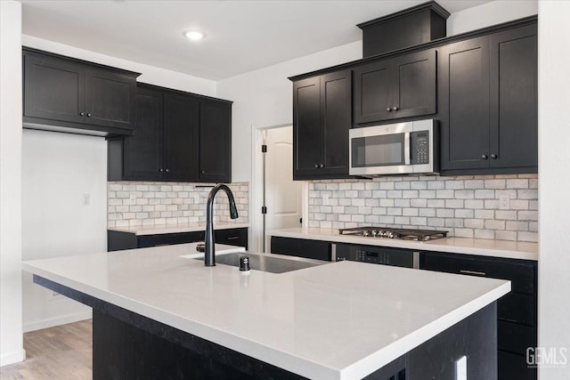 kitchen with sink, appliances with stainless steel finishes, an island with sink, light hardwood / wood-style floors, and decorative backsplash