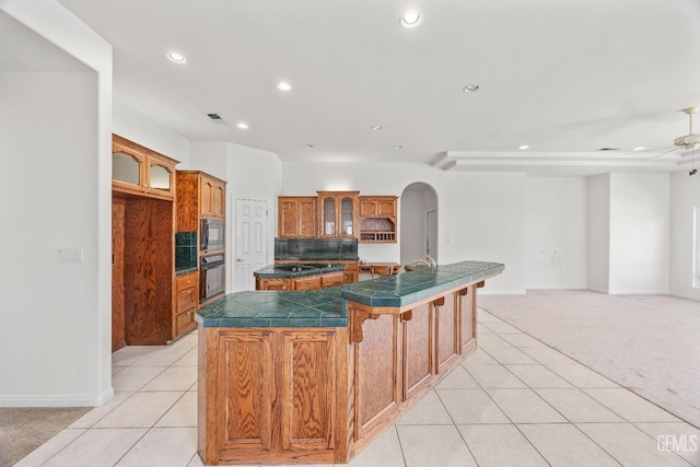 kitchen featuring light tile patterned floors, light colored carpet, brown cabinets, black appliances, and an island with sink