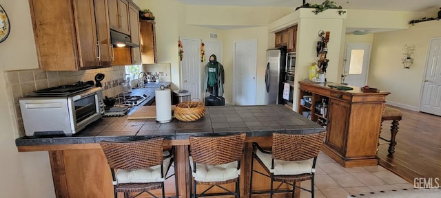 5308 Benevento Ct, Bakersfield, CA 93308 - kitchen featuring tasteful backsplash, brown cabinets, a breakfast bar area, under cabinet range hood, and freestanding refrigerator