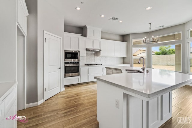 kitchen with appliances with stainless steel finishes, white cabinetry, sink, hanging light fixtures, and a kitchen island with sink