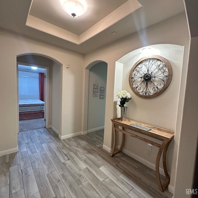 hallway featuring arched walkways, baseboards, wood finished floors, and a raised ceiling