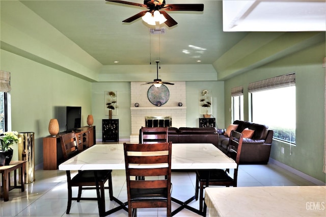 dining room with a raised ceiling, light tile patterned floors, ceiling fan, and a brick fireplace