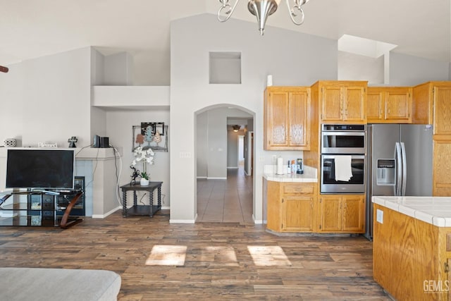 kitchen featuring tile countertops, dark wood-style floors, appliances with stainless steel finishes, arched walkways, and high vaulted ceiling