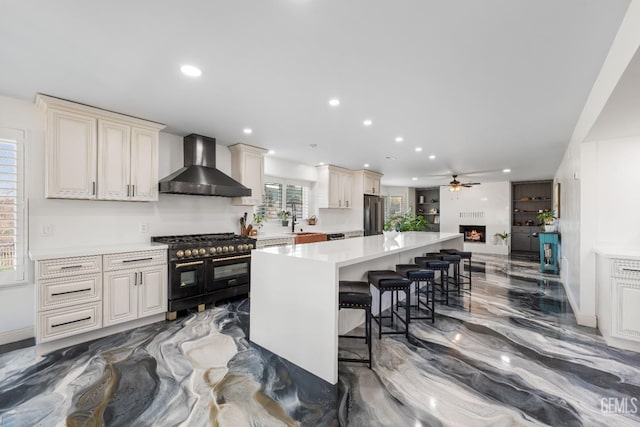 kitchen with a ceiling fan, a kitchen bar, range with two ovens, freestanding refrigerator, and wall chimney exhaust hood